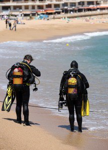 Two scuba divers in full gear walking into the sea from a sandy beach lined with hotels