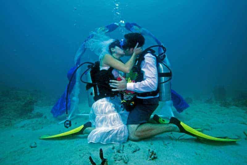 A bride and groom in scuba gear kiss underwater in front of a decorated wedding arch