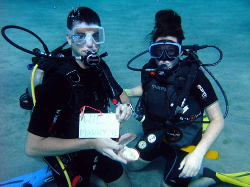 Two divers kneel underwater at Dive Grenada during a marriage proposal, one holding a slate that reads will you marry me