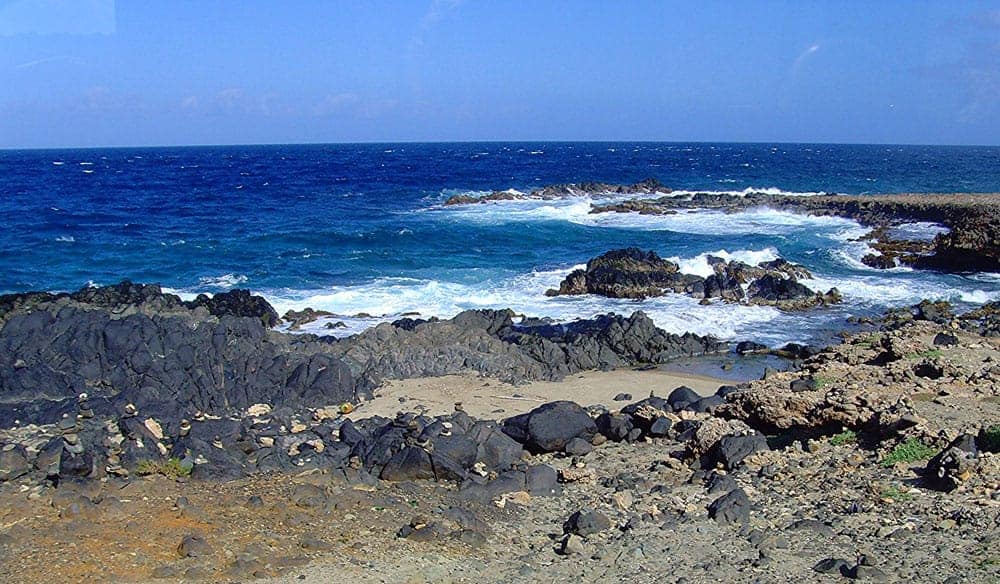 Black volcanic rock and pale sand fringing deep blue water at Black Beach on the Aruba coast