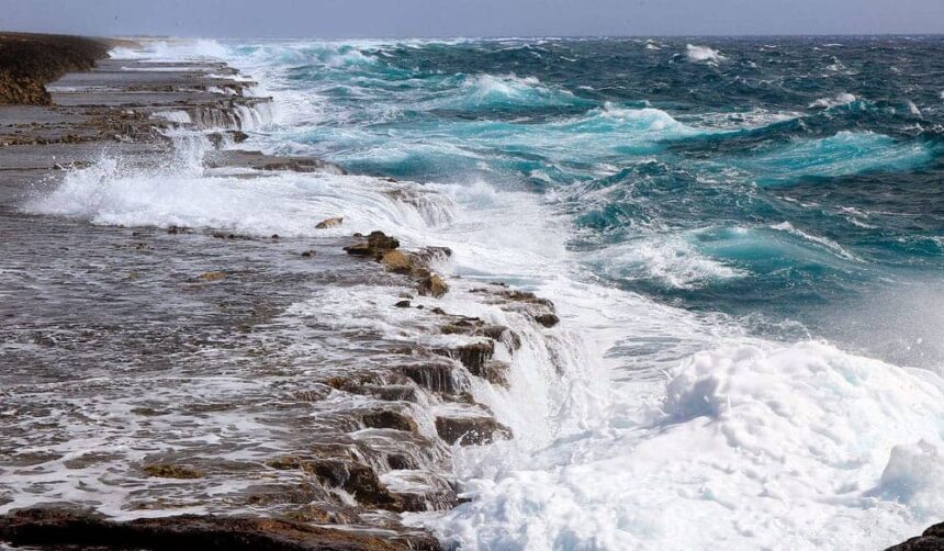 Waves breaking hard over a flat rocky shelf into churning turquoise water at Punta Basora, Aruba