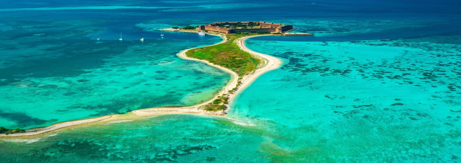 Aerial view of a small Florida island with a historic brick fort, a curving sandbar and turquoise shallows