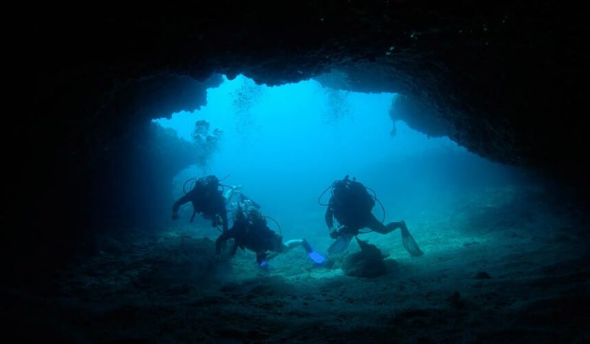 Three scuba divers silhouetted at the mouth of a dark underwater cave in Guadeloupe