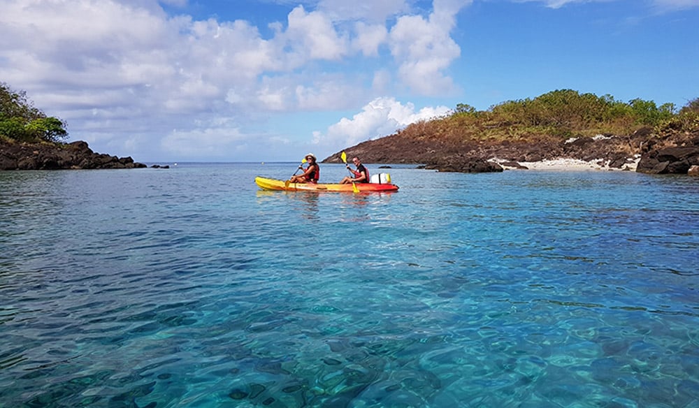 Two kayakers paddling across clear turquoise water beside a rocky green headland at Point Malendure