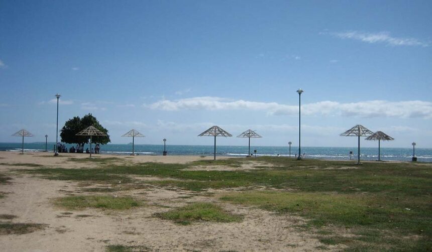 Sandy shoreline park with metal shade shelters and light surf under a bright blue sky