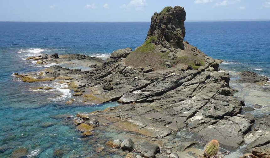 Jagged rock pinnacle rising from a low rocky islet with clear shallow water all around