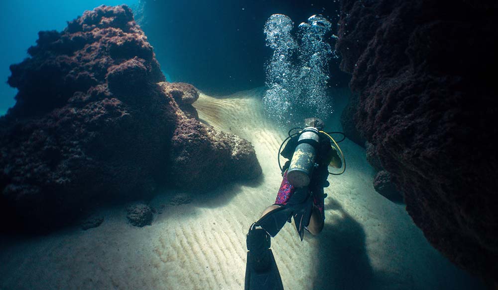 Scuba diver finning along a sandy channel between two dark rock walls