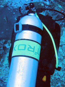 Close-up underwater of a scuba tank labeled Nitrox strapped to a diver's BCD