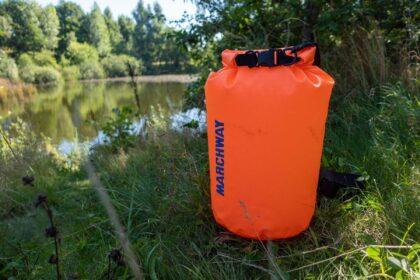 MARCHWAY orange floating dry bag rolled shut and standing in tall grass beside a calm lake