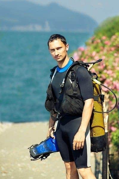 diver standing on a lakeside path with scuba gear and pink flowers behind him