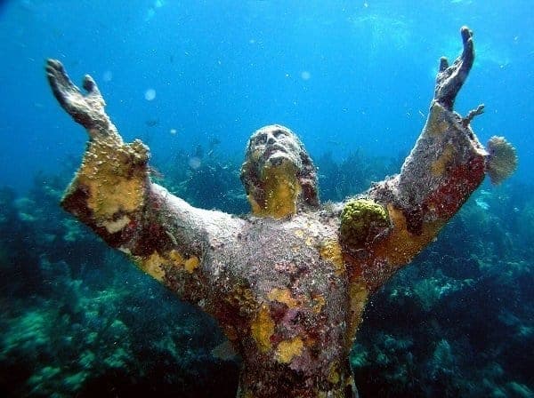 A coral-encrusted underwater statue with arms raised toward the surface