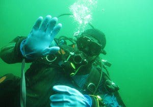 A diver gives a hand signal in murky green water typical of cold-water diving