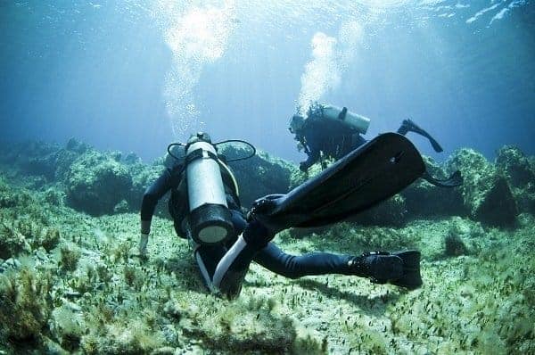 Diver glides low over a shallow reef while another diver follows behind