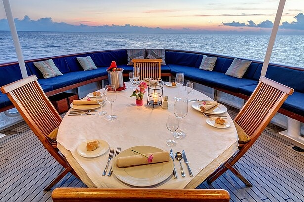 Round dinner table set on the Pelagian's aft deck with wine glasses and a pastel sunset sky