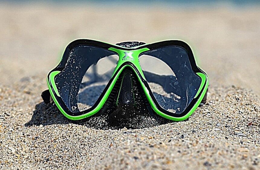 Mares X-Vision mask half buried in soft beach sand with the shoreline blurred behind