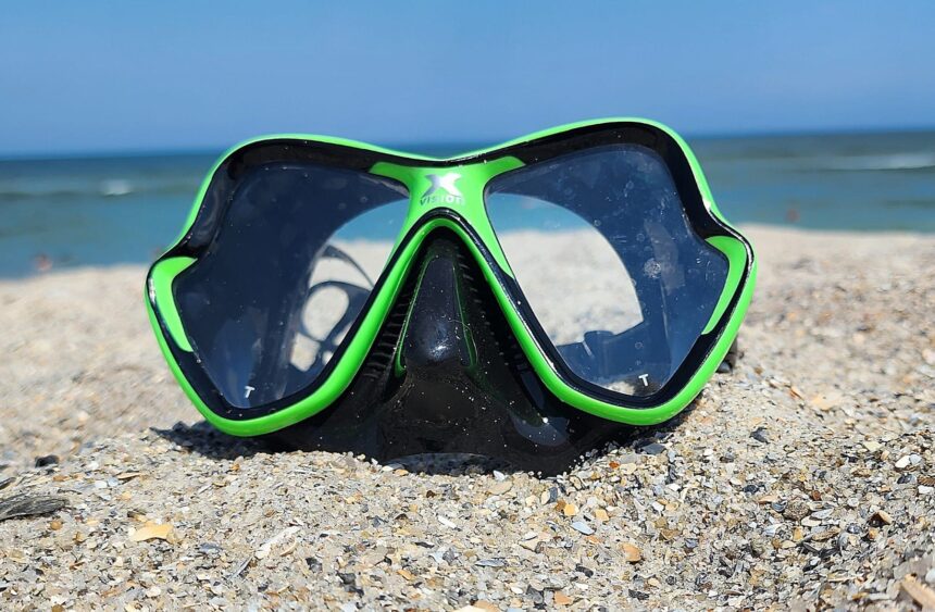 Mares X-Vision mask standing in shell strewn sand with the sea and blue sky behind