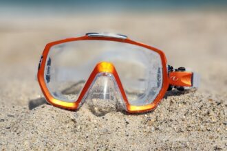 TUSA Freedom Elite mask with an orange frame and clear skirt propped upright in beach sand