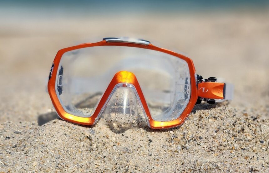 TUSA Freedom Elite Dive Mask TUSA Freedom Elite mask with an orange frame and clear skirt propped upright in beach sand