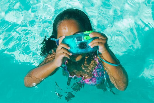 Young girl in a bright turquoise pool holding a compact blue waterproof camera up to her face