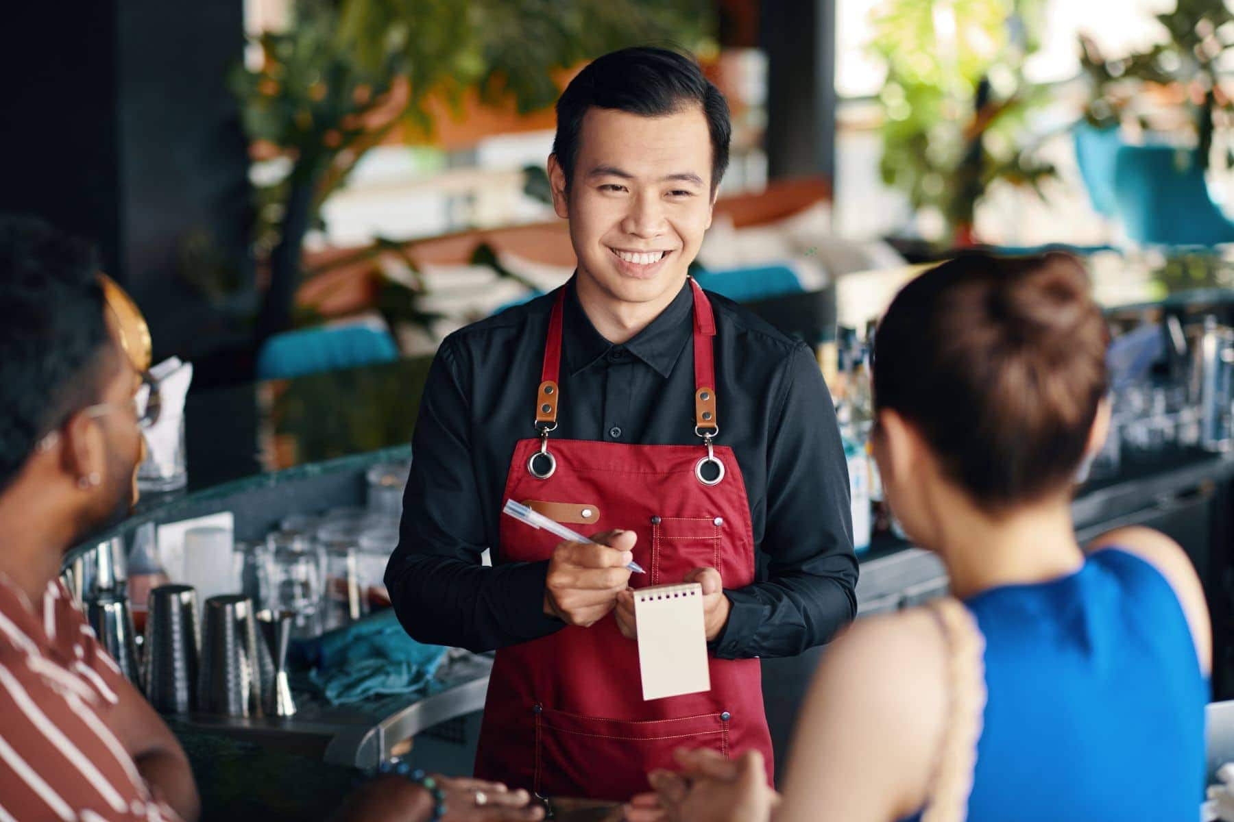 A waiter in a red apron takes an order from two smiling guests at a bar counter