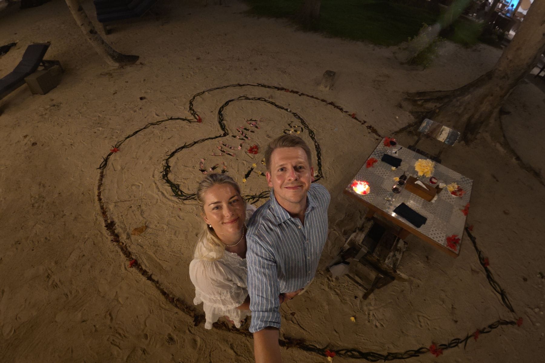 Couple embracing inside a heart shaped design drawn in the sand for a beach dinner setup