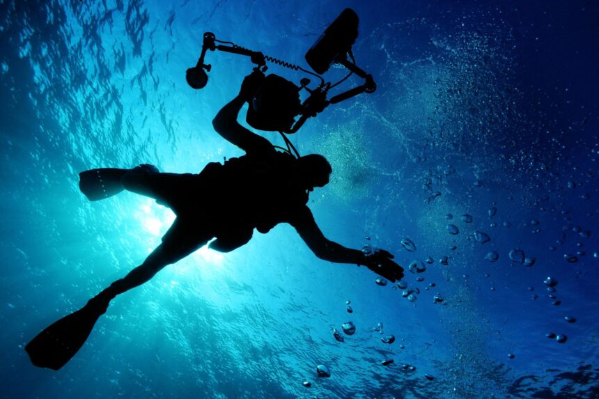 Silhouette of a scuba diver with a camera rig ascending through blue water toward sunlit surface