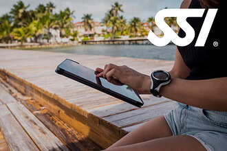 A woman wearing a dive watch works on a tablet while sitting on a wooden dock beside a tropical waterfront