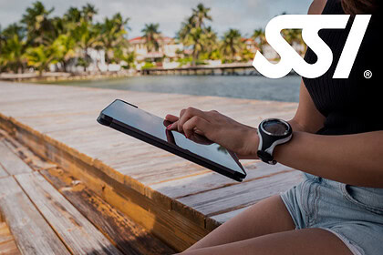 A woman wearing a dive watch works on a tablet while sitting on a wooden dock beside a tropical waterfront