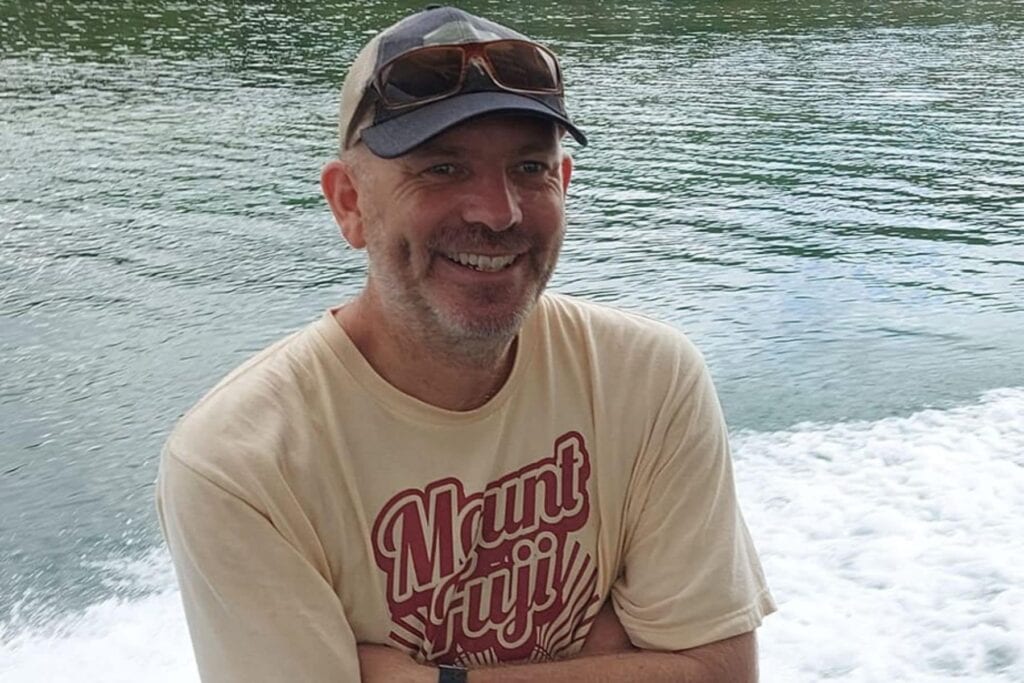 Smiling bearded man in a cap with sunglasses on top, wearing a Mount Fuji T-shirt on a boat
