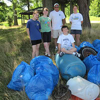 People standing beside filled blue rubbish bags, a barrel and a tire under trees