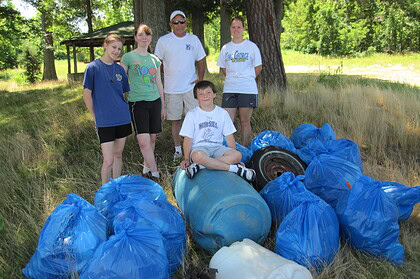 People standing beside filled blue rubbish bags, a barrel and a tire under trees