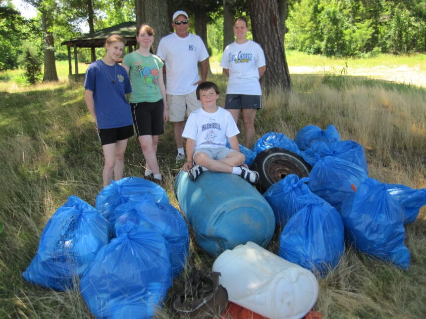 People standing beside filled blue rubbish bags, a barrel and a tire under trees